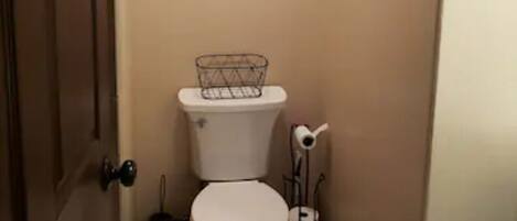 A narrow bathroom with a toilet, a decorative wall piece above it, and a trash can beside the toilet.