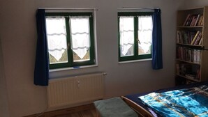 Ein Schlafzimmer mit zwei Fenstern, einem Heizkörper, einem Bett mit blauer und gelber Bettdecke und einem Bücherregal mit Büchern.