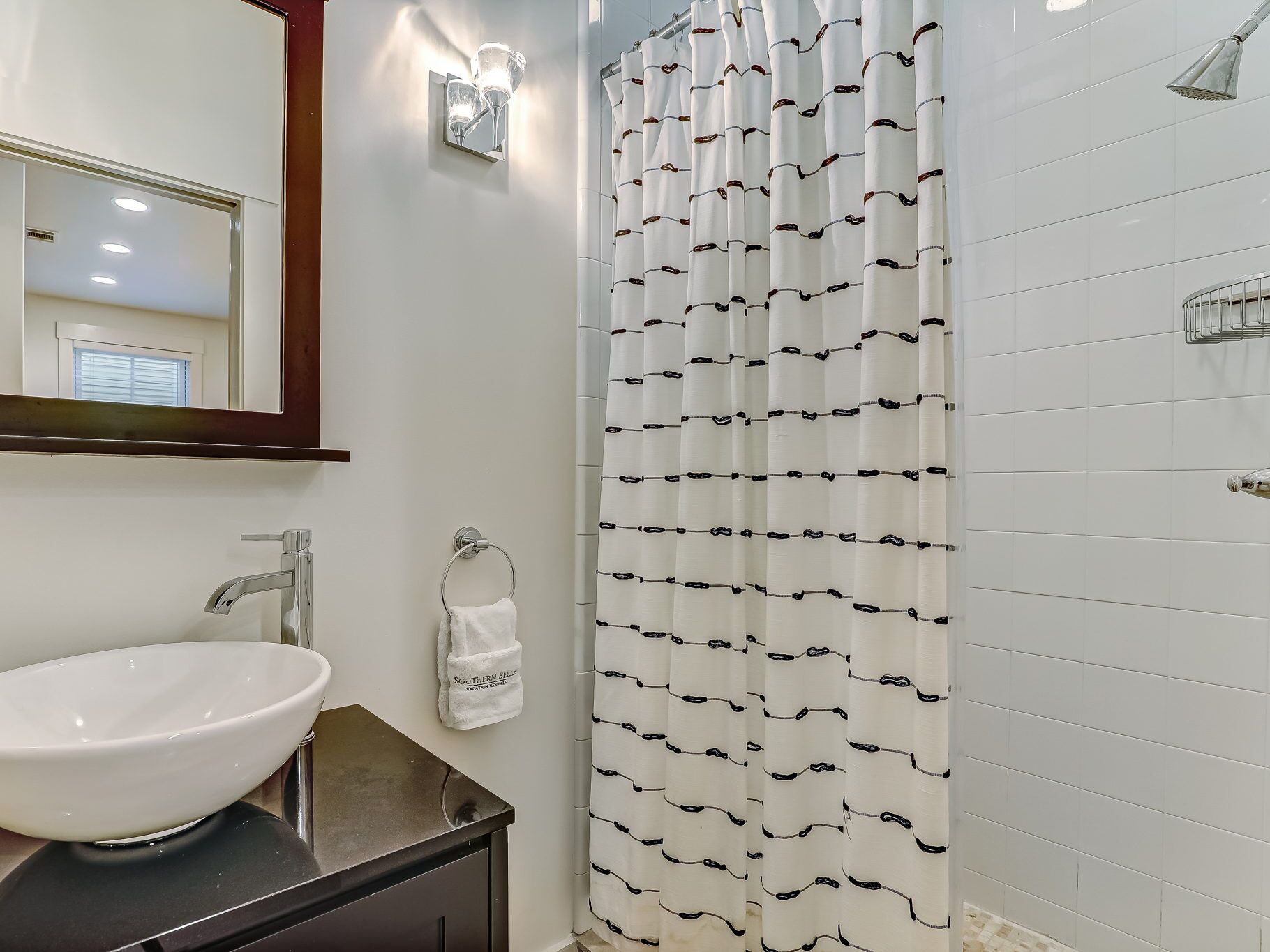 Une salle de bain avec un lavabo blanc, un porte-serviettes, un rideau de douche à motifs et un miroir agrémenté d’appliques murales.