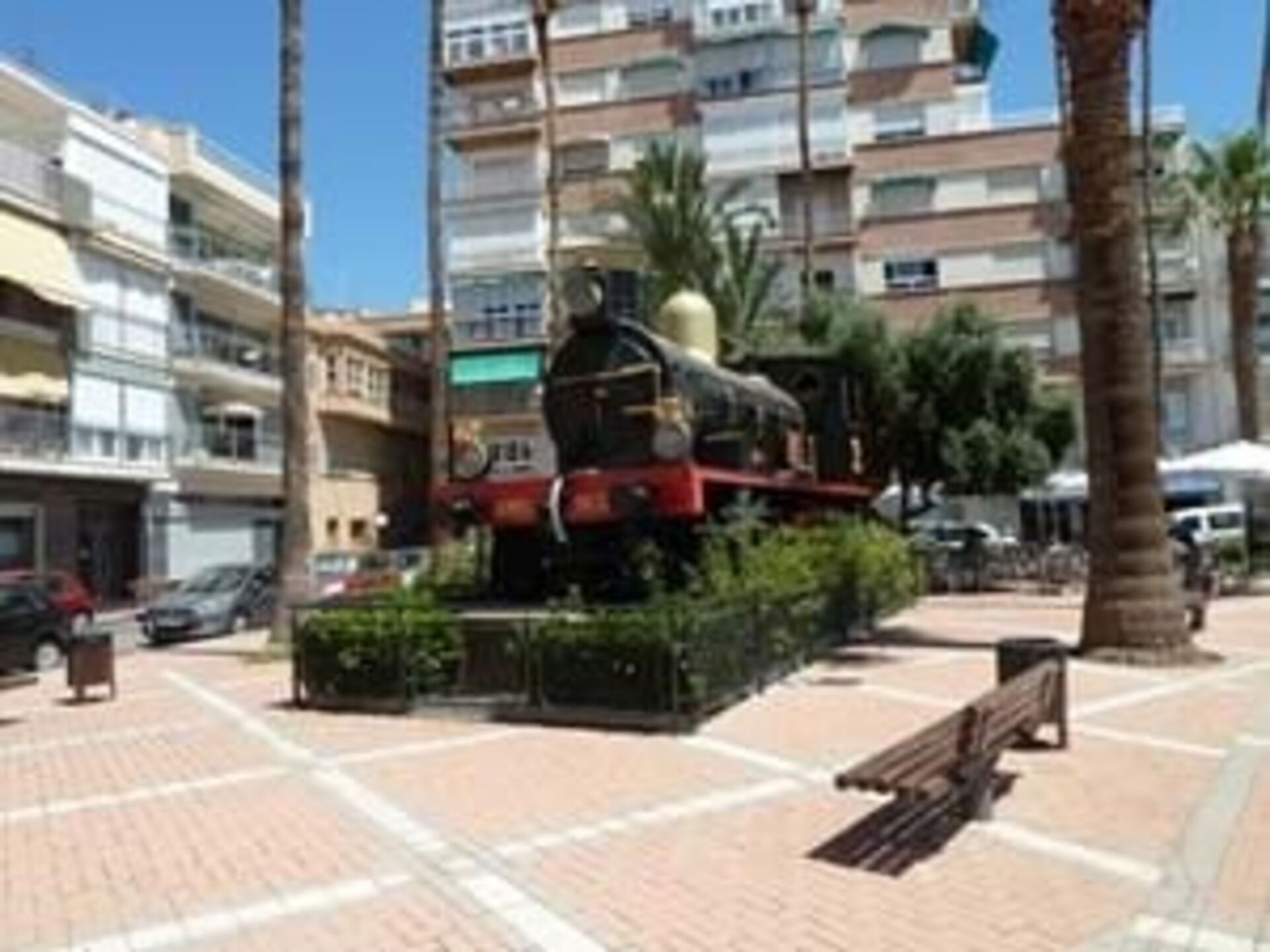 A train statue in a public square with surrounding buildings and palm trees.