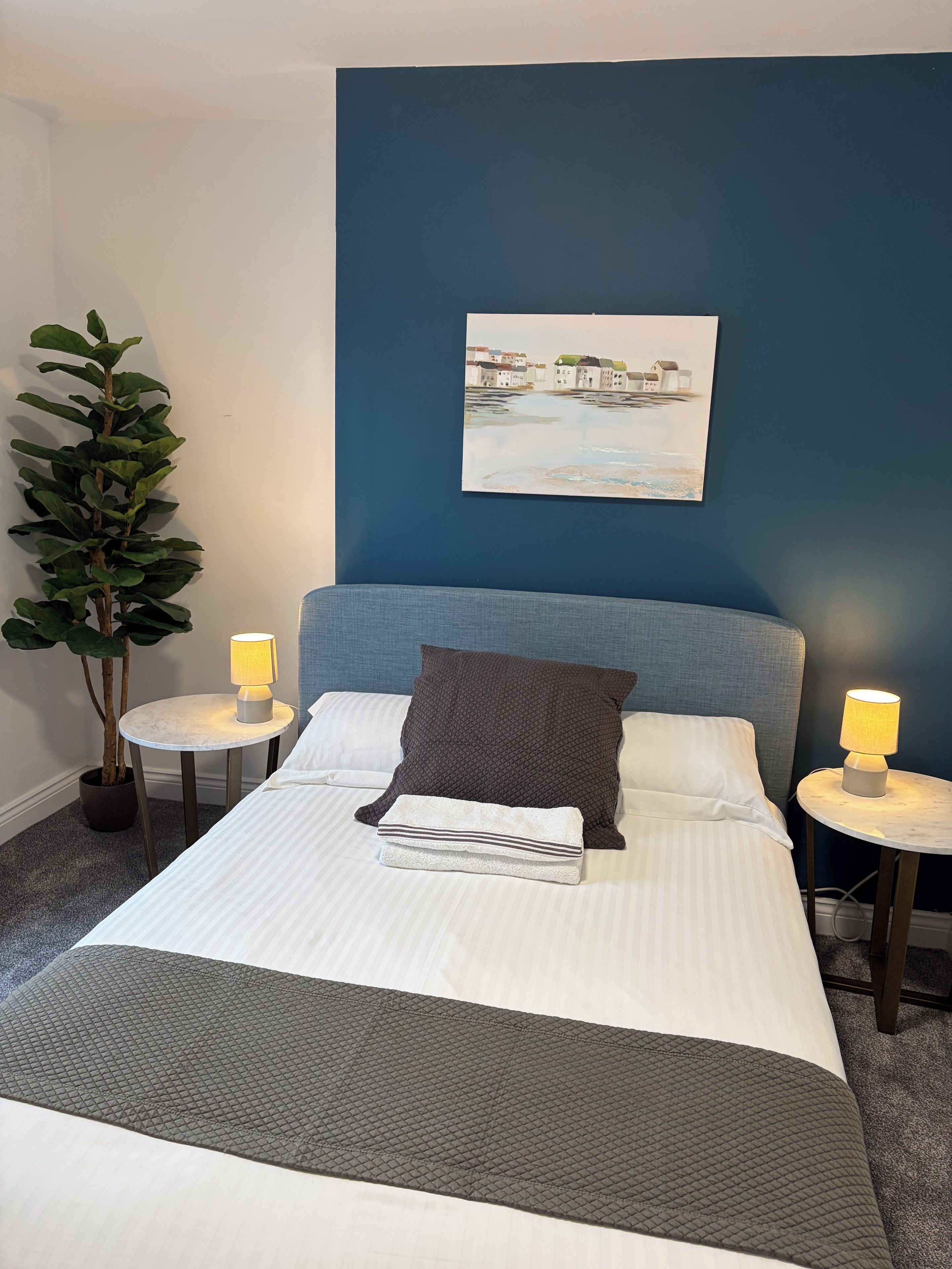 A neatly made bed with pillows and a blanket, two bedside tables with lamps, a wall-mounted painting, and a potted plant.
