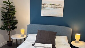 A neatly made bed with pillows and a blanket, two bedside tables with lamps, a wall-mounted painting, and a potted plant.