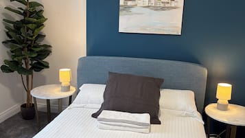 A neatly made bed with pillows and a blanket, two bedside tables with lamps, a wall-mounted painting, and a potted plant.