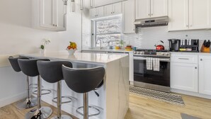 A modern kitchen with white cabinets, a central island with bar stools, stainless steel appliances, and a large window providing natural light. - LUXE 3BD/2BA 29 min>NYC w/Parking & deck  (Hastings-on-Hudson)