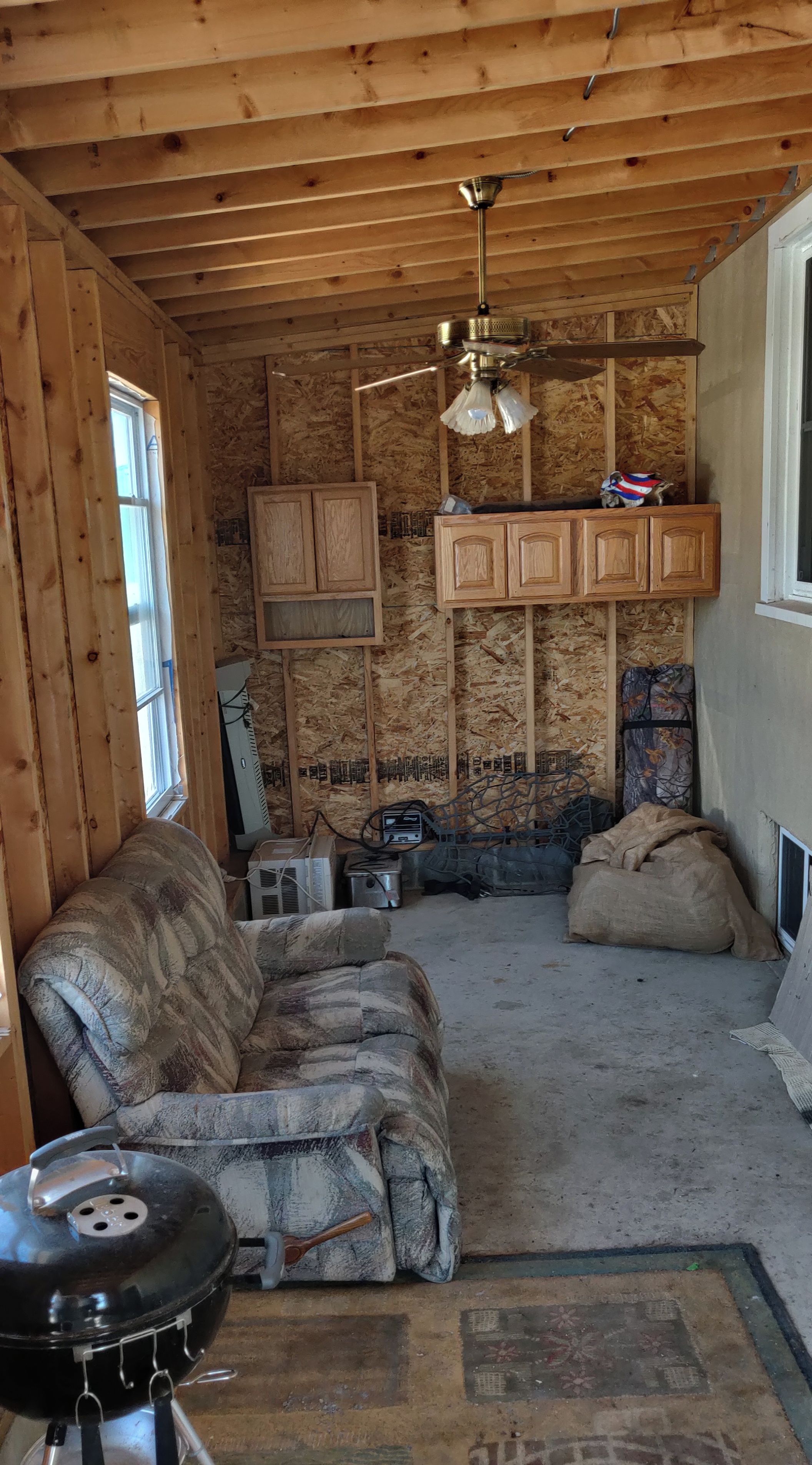 A room with wooden walls and ceiling, a ceiling fan, a couch, a grill, and a window.