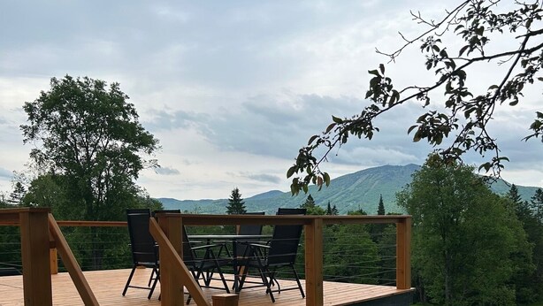 Une terrasse en bois, agrémentée d’une table et de chaises, entourée d’une rambarde en métal, et offrant une vue sur une zone herbeuse et des collines lointaines.