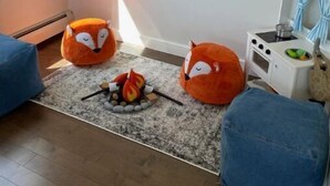 Un salon chaleureux avec une cheminée, deux fauteuils en forme de renard orange, un pouf bleu et un poêle blanc.