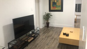 A living room with a flat-screen TV on a glass stand, a potted plant, a framed abstract painting, and a wooden table with a few items on it. - Lovely 4 bedroom Home with a patio (Oakland)