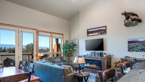 A spacious living room with a large sofa, a dining table, and a fireplace. There is a television mounted on the wall and a mounted animal head decoration. - 4 king beds w/ ensuites | Double the Living Space! (Bigfork)