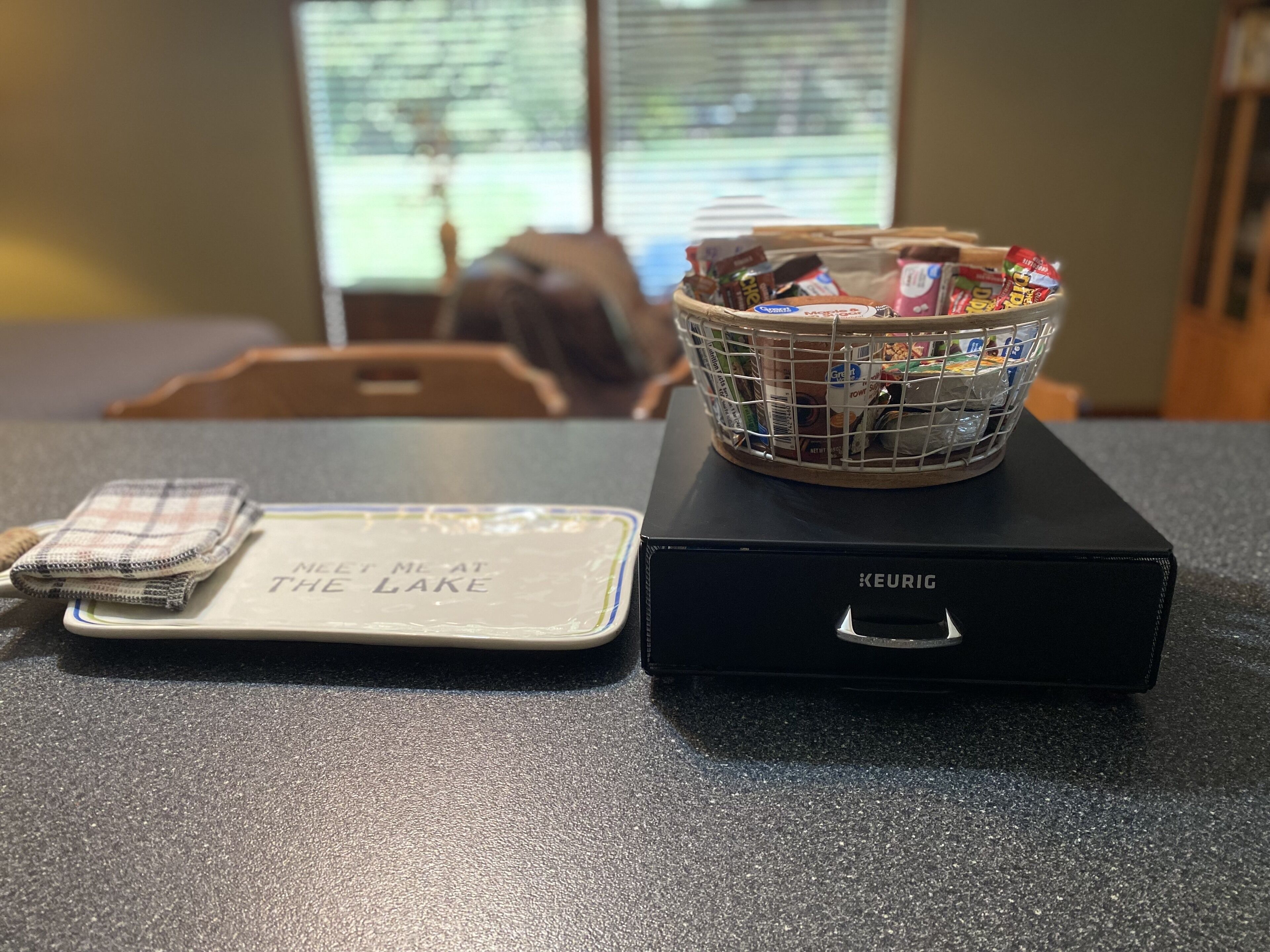 Eine Küchenarbeitsplatte mit einer Keurig-Kaffeemaschine, einer Snackkorb und einer Unterlage mit dem Aufdruck 'Meet Me at the Lake'.