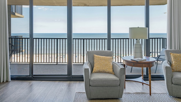 Uma varanda com vista para a praia, dois poltronas com almofadas estampadas, uma mesa redonda pequena com abajur e piso de madeira.