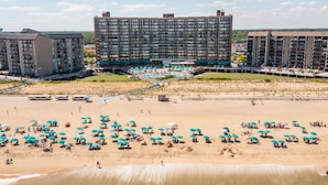 Um resort à beira-mar com vários edifícios, uma praia arenosa com muitos guarda-sóis e uma área de piscina.