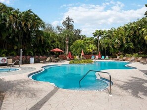 A swimming pool with a curved edge, surrounded by lounge chairs and umbrellas, with palm trees in the background. - Newly remodeled coach home & golf membership in Vasari Country Club. (Bonita Springs)