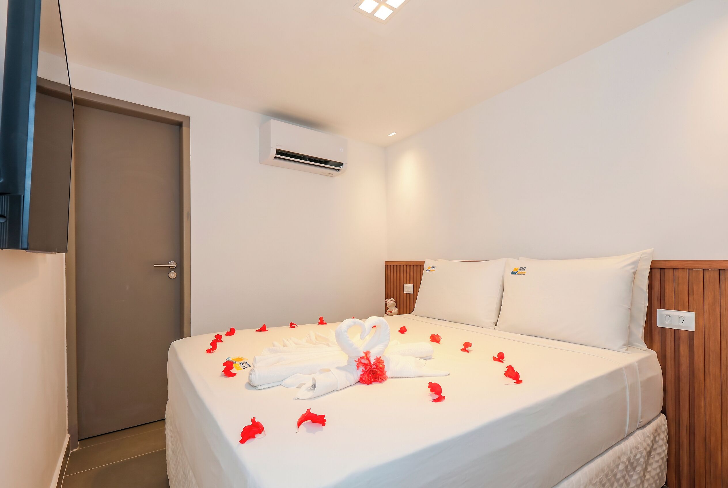 Ein Hotelzimmer mit Bett, weißer Bettwäsche, Holz-Kopfteil und einer Klimaanlage sowie einer Tür.
