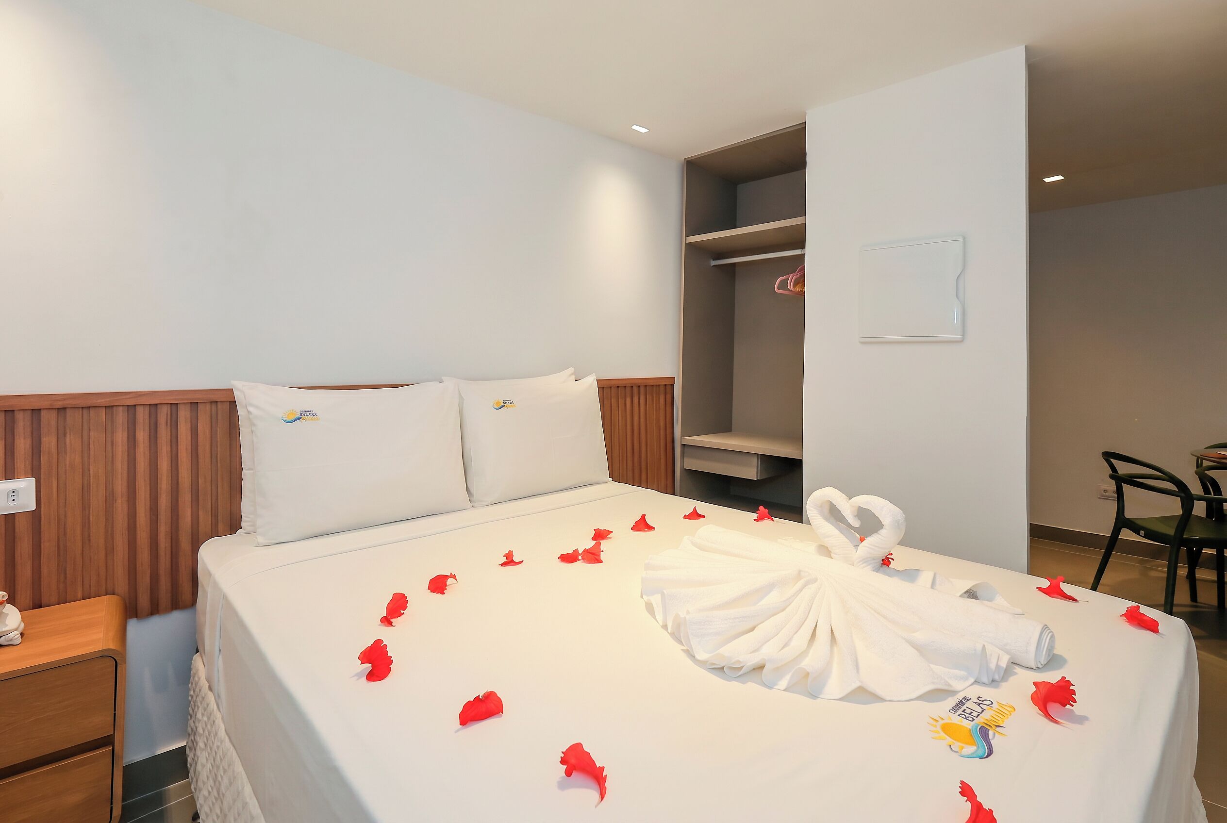 A hotel room with a neatly made bed, a wooden headboard, a wardrobe, and a small table with a chair.