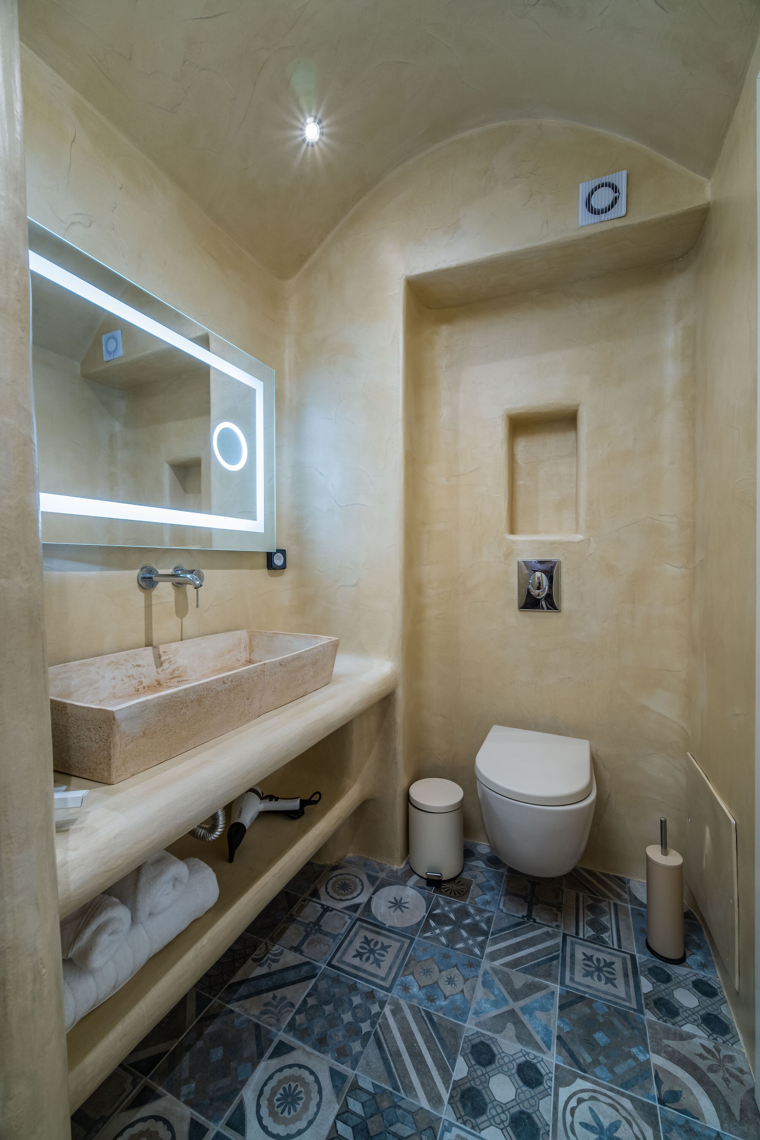 Un baño con pared de azulejos color beige, un lavabo con espejo, un inodoro y suelo con estampado.