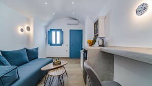 A compact living space with a sofa, a small dining table, a kitchen area with a countertop, and a blue door. - Aegean Blue Luxury Suites (Santorini)