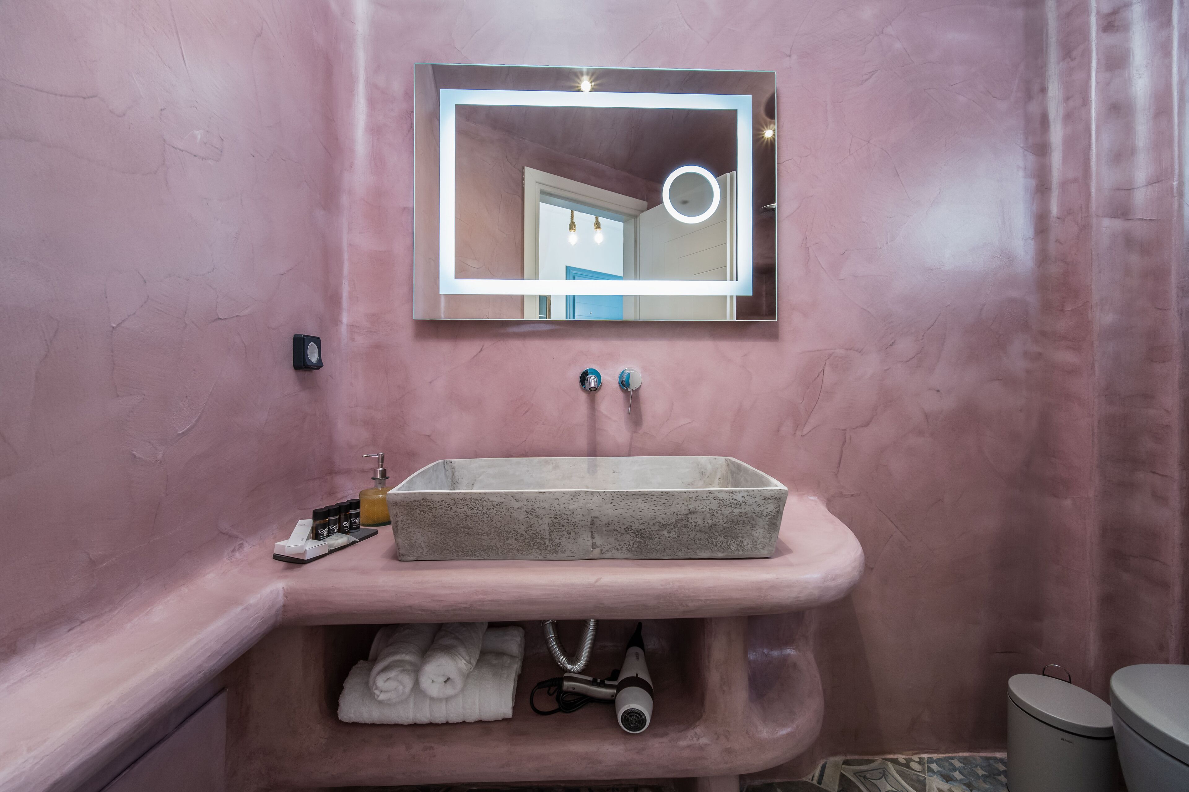 Een badkamer met een roze wand, een rechthoekige spiegel met verlichting, een rechthoekig wastafelmeubel en een handdoekhouder.