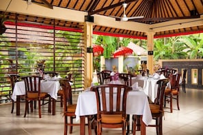 Un comedor interior con mesas dispuestas para los huéspedes, sillas de madera y vistas a un jardín tropical.