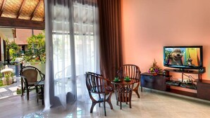 A living room with a TV, wooden furniture, and a view of a garden. - Rossa Garden Hotel (Karangasem)
