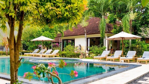 A pool area with lounge chairs and umbrellas, surrounded by lush greenery and a building with a tiled roof. - Rossa Garden Hotel (Karangasem)