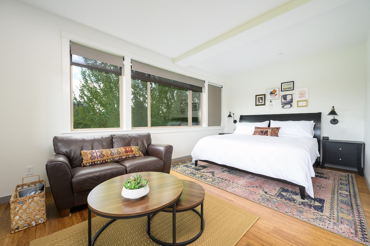 A bedroom with a large bed, a brown leather sofa, a round coffee table with a plant, and a window with a view of trees.