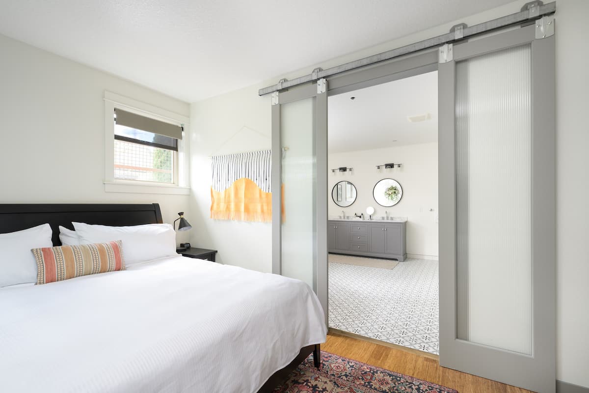A bedroom with a bed, a nightstand, a window with blinds, a bathroom with a mirror, and a sliding door to another room.