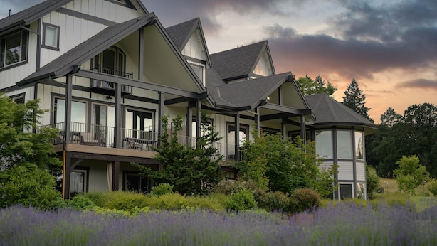 Une maison moderne à deux étages, dotée d’un balcon, entourée d’une végétation luxuriante et d’un champ de fleurs violettes.