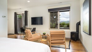 Une chambre d’hôtel moderne dotée d’un grand lit, de deux fauteuils, d’une petite table avec un bol, d’un téléviseur à écran plat, d’une cheminée et d’une grande fenêtre donnant sur un paysage verdoyant.