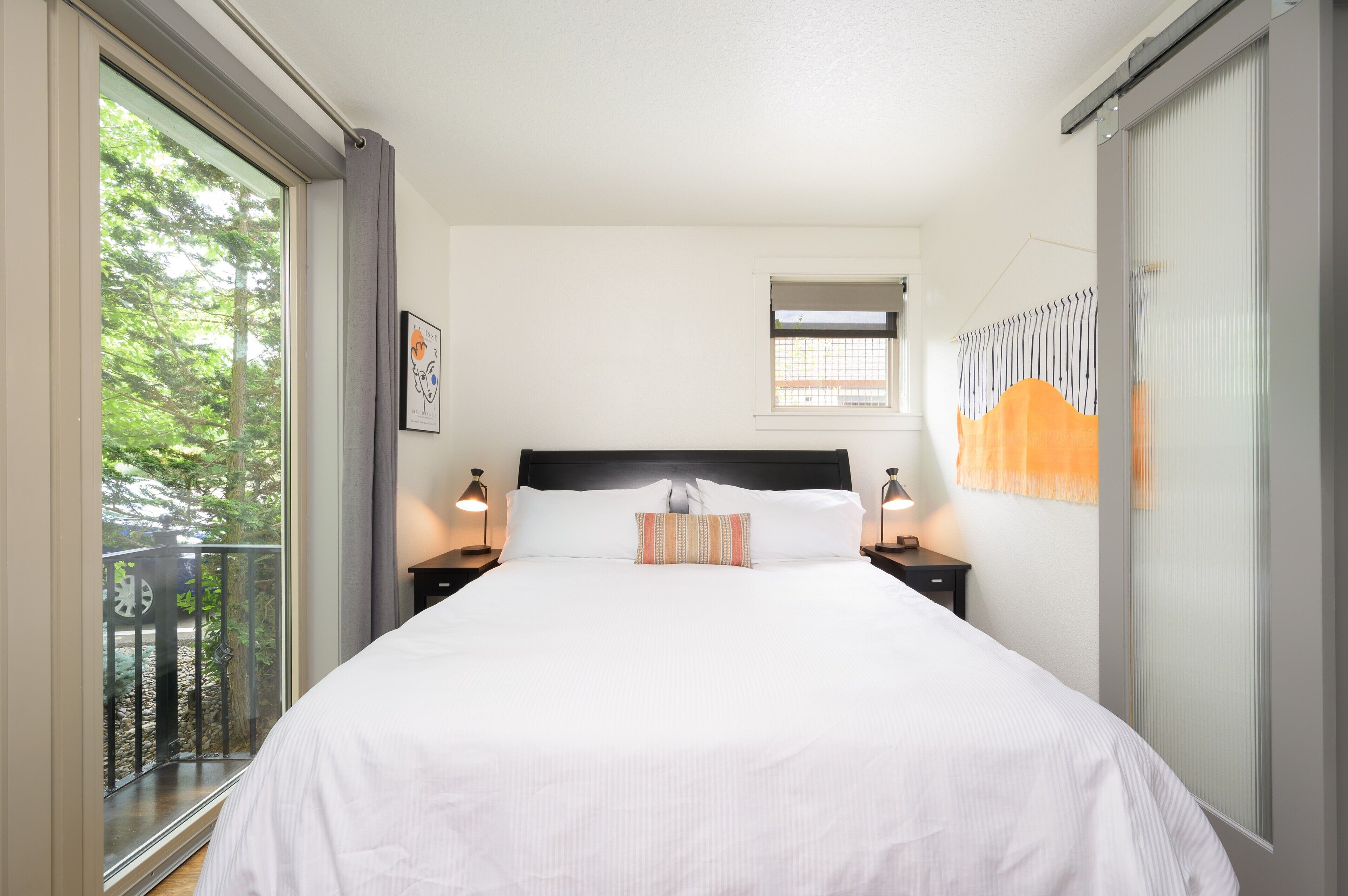 Een net opgemaakt bed met een kussen, een klein raam met uitzicht op bomen, en een balkon met een hekwerk.