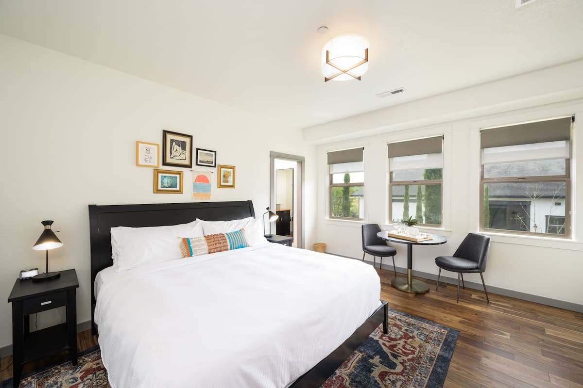 A hotel room with a large bed, a desk with a chair, and a small table with two chairs. There are framed pictures on the wall and a window with a view of trees.