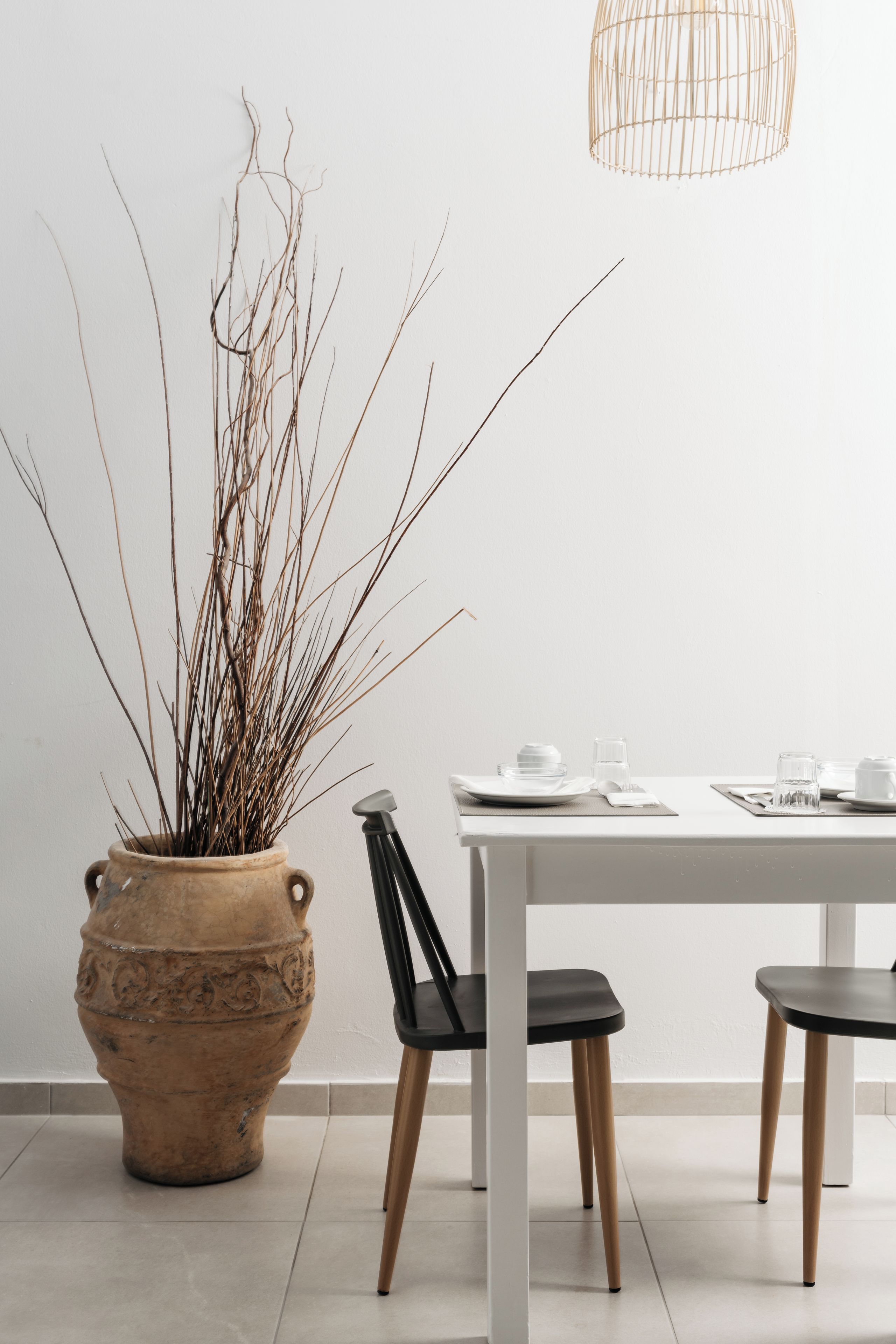 A dining area with a white table, black chairs, a terracotta pot with branches, and a hanging light fixture.