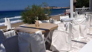 A beachside restaurant with white-covered tables and chairs, a wooden table in the foreground, and a view of the ocean and beach in the background. - Petradi Beach Lounge Hotel by GHH (Rethymno)