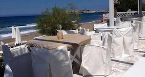 Un restaurante frente al mar con mesas y sillas blancas, una mesa de madera en primer plano, y vista al océano y la playa al fondo.