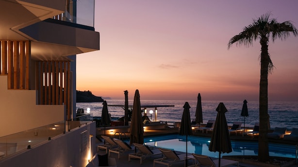 A rooftop pool area with lounge chairs and umbrellas, overlooking the sea at sunset.