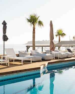 Zona de piscina con sillones de descanso y sombrillas, un muelle de madera y vista al mar.