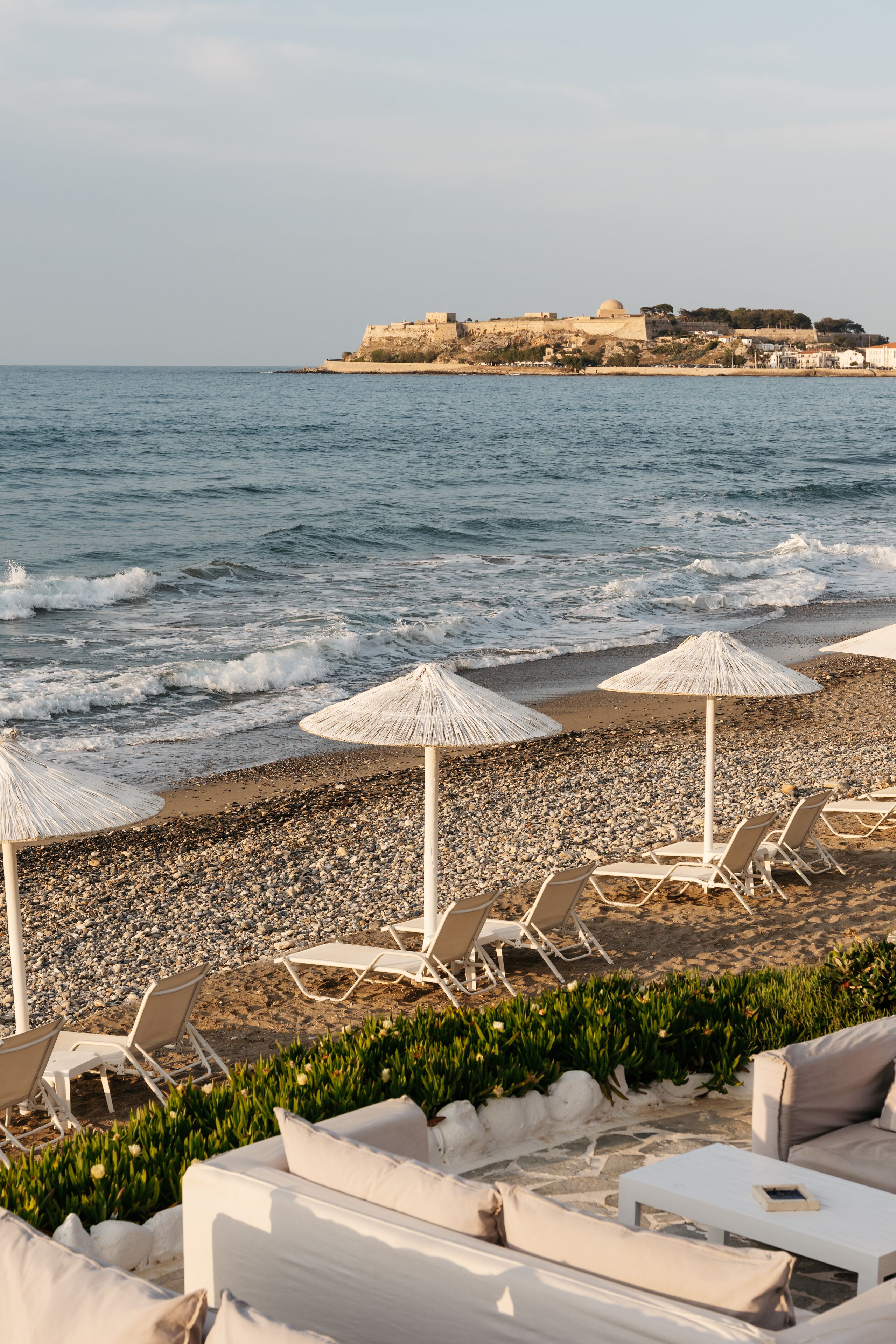 A beach with white lounge chairs and umbrellas, a rocky shoreline, and a distant view of a fortified structure.