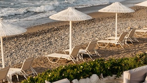 A beach with white lounge chairs and umbrellas, a rocky shoreline, and a distant view of a fortified structure. - Petradi Beach Lounge Hotel by GHH (Rethymno)