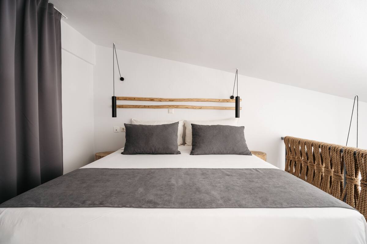 Een moderne slaapkamer met een houten hoofdbord, twee kussens, een grijze deken en een rieten stoel.