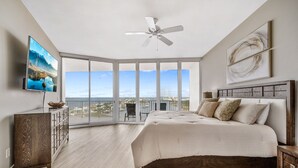 A bedroom with a large bed, a ceiling fan, a TV, and a balcony with a city view.