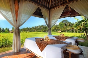 Quarto com duas camas, vista para um campo verde e estrutura com telhado de palha.