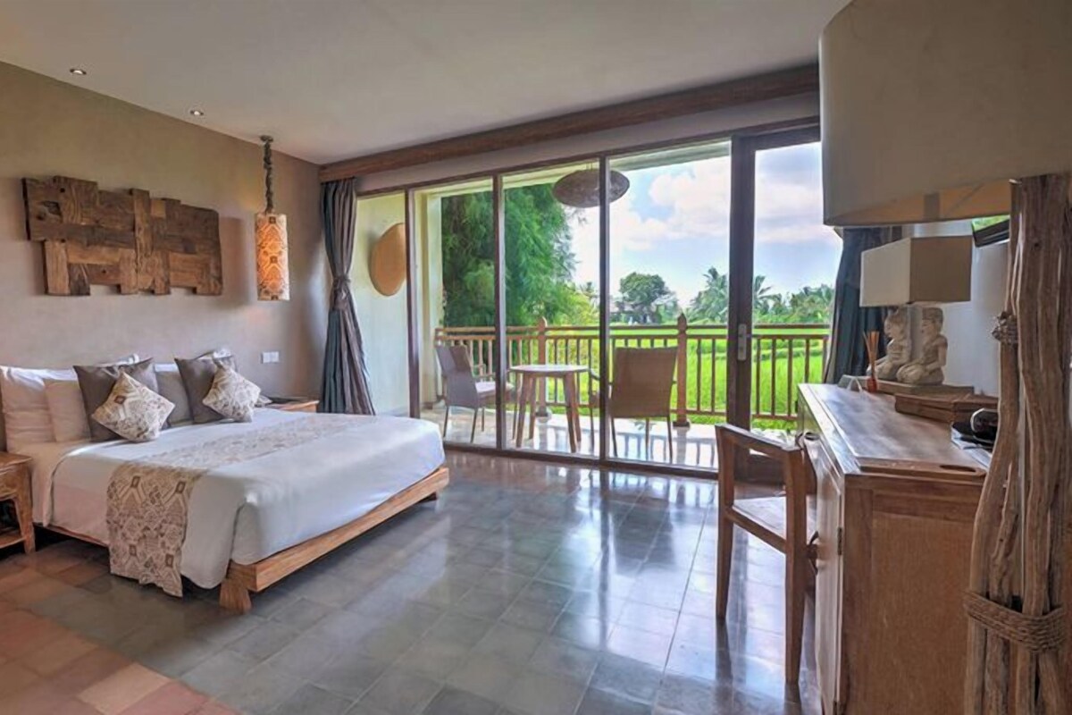 Quarto com cama grande, mesa de cabeceira, escrivaninha e varanda com vista para o verde.