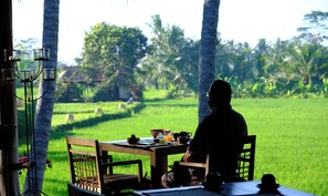 A dining table with chairs, a candle holder, and a view of a lush green field and trees. - Mathis Retreat Ubud (Ubud)