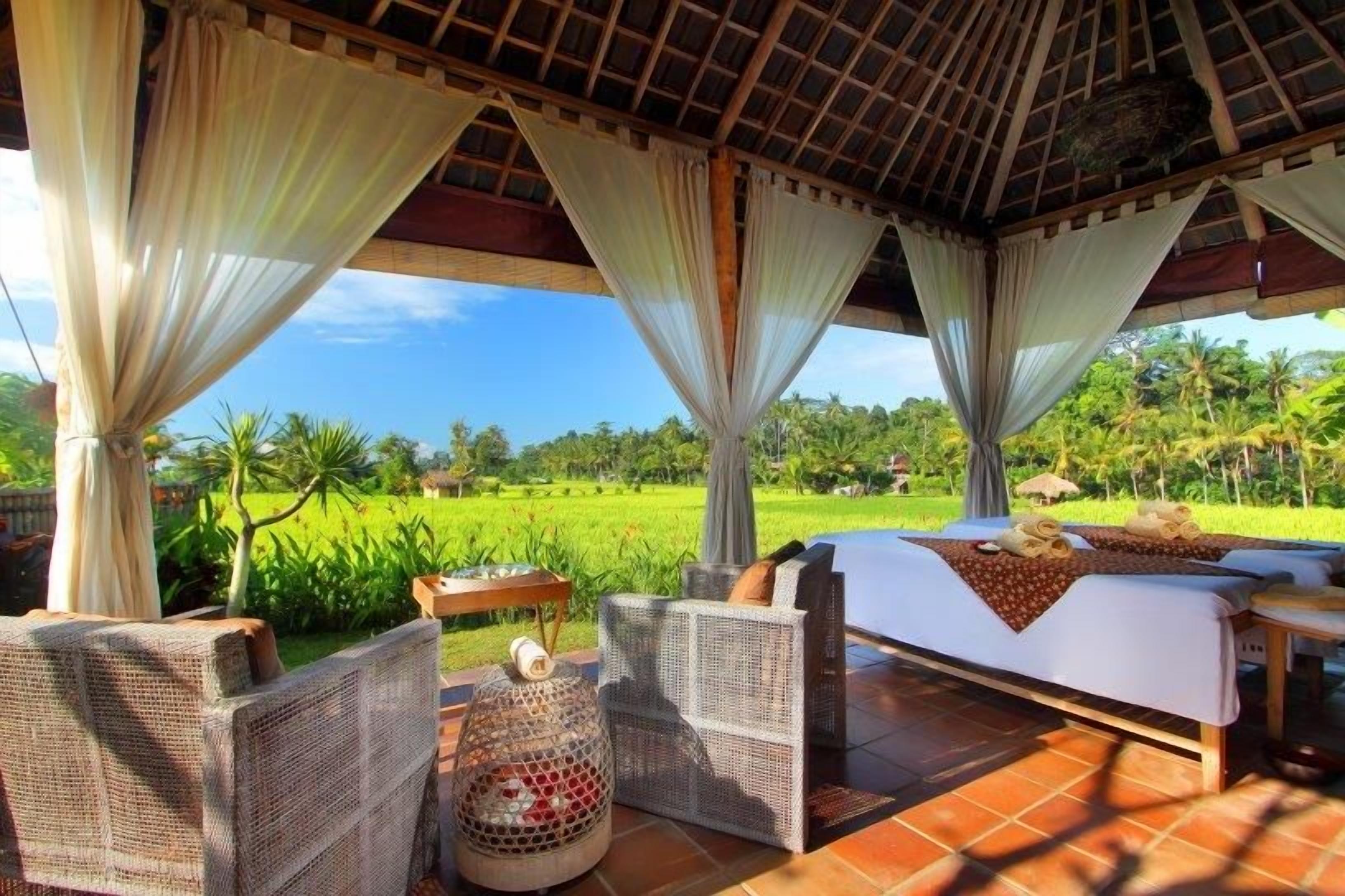 Un área de estar techada al aire libre con una cama, muebles de mimbre y vistas a un campo verde exuberante.