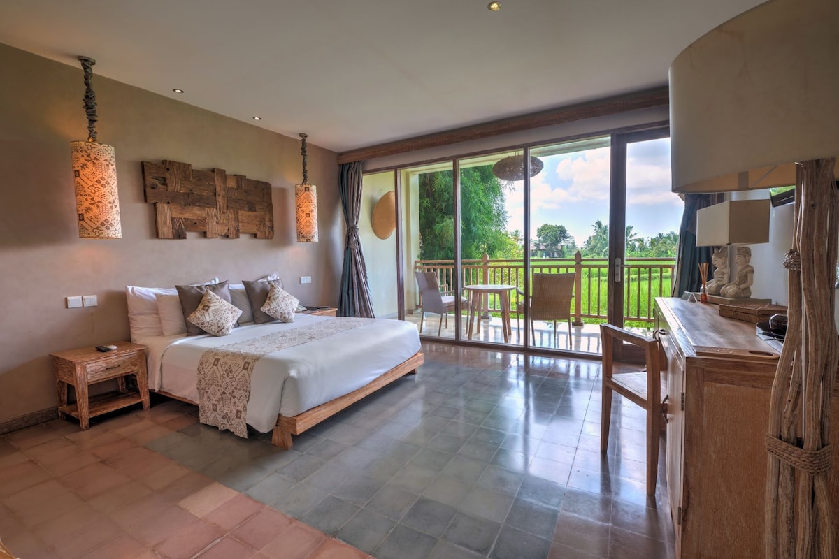 Quarto espaçoso com uma cama grande, uma mesa de cabeceira de madeira, uma escrivaninha e uma varanda com vista para o verde.