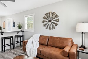 A living room with a brown leather sofa, a white throw blanket, a round wall decoration, a lamp, and a window with blinds.