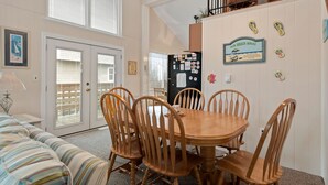 A dining area with a wooden table and chairs, a striped couch, and a kitchen with a refrigerator and decorative items. - Sun Hut: Oceanside Home w/ Private Pool, Short Walk to the Beach! (Corolla)