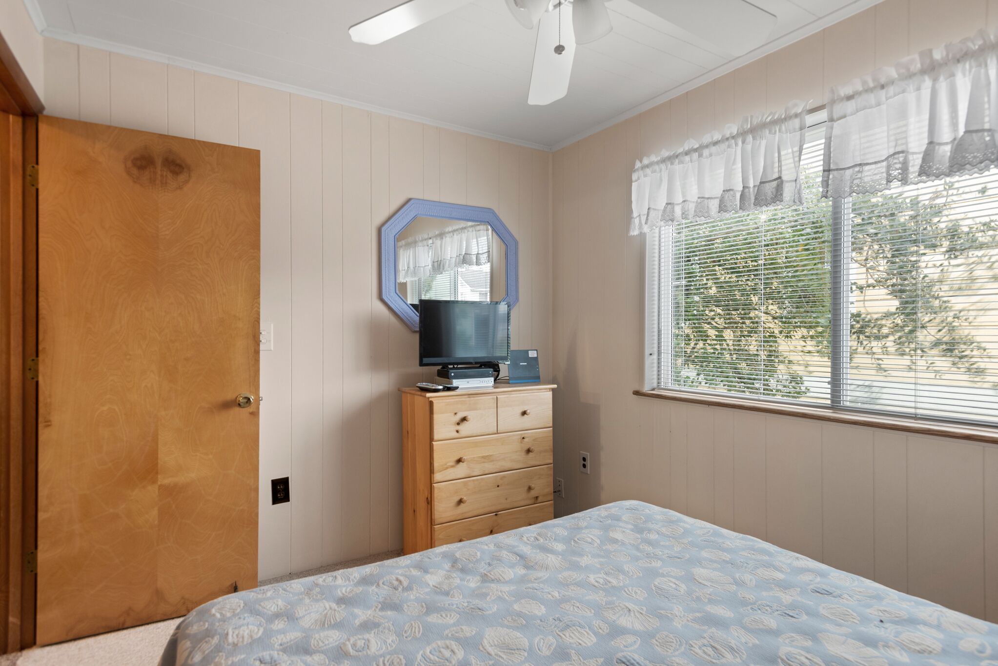 A bedroom with a bed, wooden dresser, television, and a window with blinds.