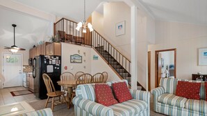 A cozy living room with striped sofas, a dining area with wooden chairs, a kitchen with a refrigerator, and a staircase leading to an upper level. - Sun Hut: Oceanside Home w/ Private Pool, Short Walk to the Beach! (Corolla)