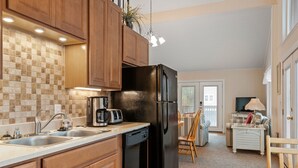 A kitchen with wooden cabinets, a black refrigerator, a coffee maker, and a dishwasher. There is a dining area with a table and chairs. - Sun Hut: Oceanside Home w/ Private Pool, Short Walk to the Beach! (Corolla)